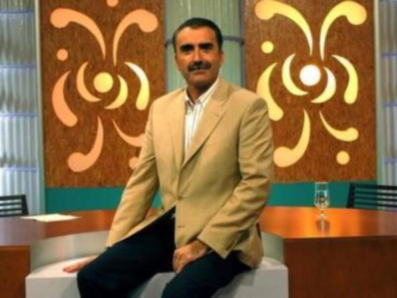 Un hombre con barba y bigote, vestido con un traje beige, se sienta en una mesa de estudio. El fondo muestra un diseño abstracto con patrones geométricos y colores vibrantes, típico de una producción televisiva.