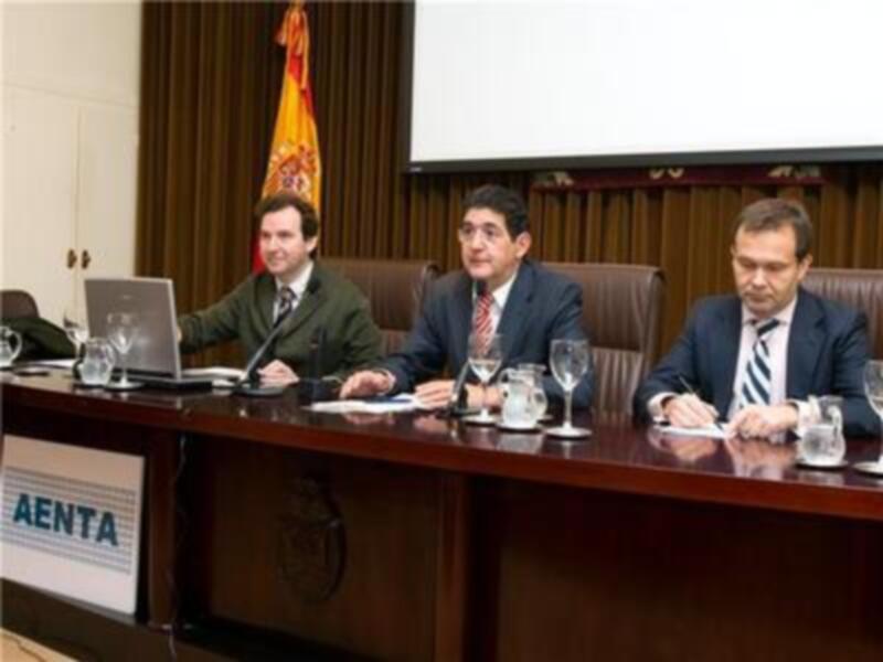 Tres hombres sentados en una mesa de conferencia, con un fondo de cortinas y una pantalla. En la mesa hay un portátil, gafas, tazones de café y un cartel con la palabra "AENTA".