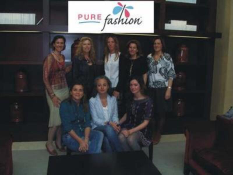 Una fila de siete mujeres posan frente a una pared con estanterías, en la parte superior se lee "PURE fashion". La imagen parece ser de una tienda o evento relacionado con la moda.
