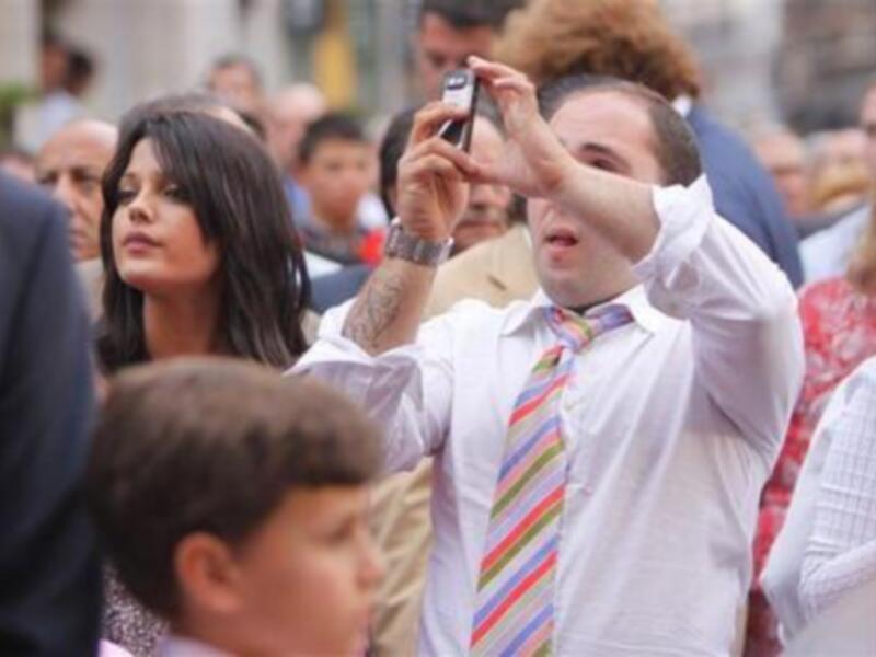 Un hombre con corbata de colores toma una foto en la calle, rodeado por personas y edificios.
