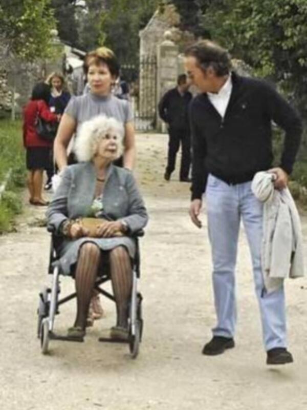 Una anciana en silla de ruedas camina con un hombre y una mujer, todos parecen estar en un entorno de jardín o parque.