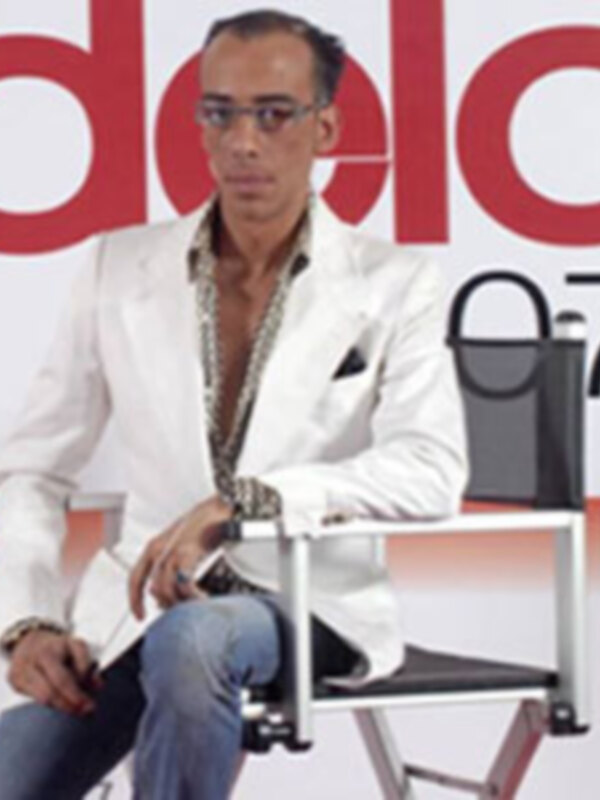 Un hombre con un traje blanco y una corbata de cristales, sentado en una silla negra con un fondo rojo y blanco.