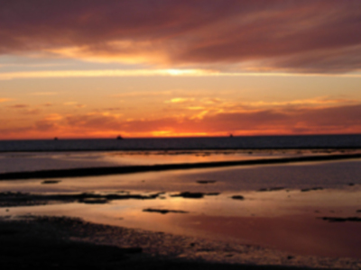 Puesta de sol sobre el mar, con tonos rojos y naranjas en el cielo. La playa está vacía, con agua lisa reflejando los colores del atardecer.