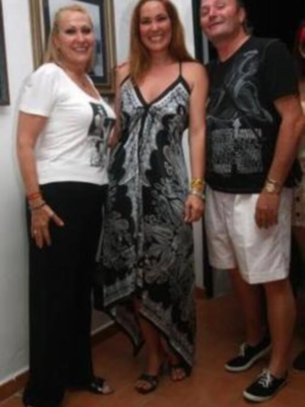 Tres personas sonrientes en una habitación con un marco de fotos en el fondo. La imagen muestra a una mujer en la izquierda, otra en el centro y un hombre en la derecha. Todos están sonriendos y parecen estar en una reunión o evento familiar.
