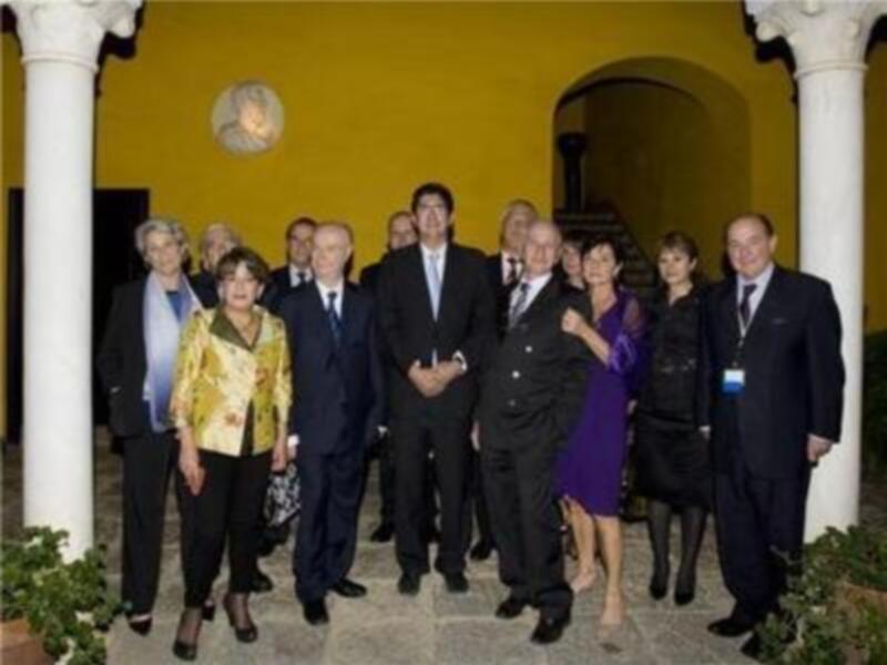 Una imagen de una reunión formal con varias personas vestidas formalmente, posando frente a un edificio amarillo con columnas blancas.