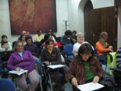Una clase en un aula con estudiantes sentados, algunas tomando notas. La imagen muestra una escena educativa en un entorno formal con carteles y sillas organizadas.