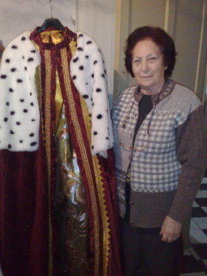A una mujer posando junto a un traje histórico. El traje tiene un chaleco blanco con puntos negros y una capa roja con detalles dorados. La mujer está vestida con un suéter a cuadros y una falda negra. La escena parece ser interior, posiblemente en un museo o exposición histórica.