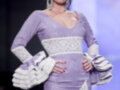 Una modelo presenta un vestido de color lila con detalles bordados blancos y mangas en volantes. La pieza se ajusta a la figura, destacando un escote profundo y mangas con volantes. La modelo lleva una corona en la cabeza, complementando el look.