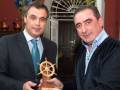 Dos hombres sostienen un trofeo en forma de rueda de timón, posiblemente en un evento de premiación o reconocimiento.