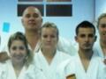 Una escena en un dojo, cuatro personas vestidas con uniformes blancos de karate posan para una foto. En el fondo, se pueden ver paneles con información técnica del entrenamiento.