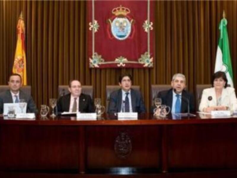 Celebración formal en una sala de conferencias con banderas nacionales y un escudo real en el fondo. Un grupo de personas se sientan frente a una mesa con documentos y copas, posiblemente en un evento político o gubernamental.