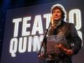 Un hombre con una camisa de cuero y un micrófono en el escenario frente a un fondo con la palabra "Teatros Quintos".