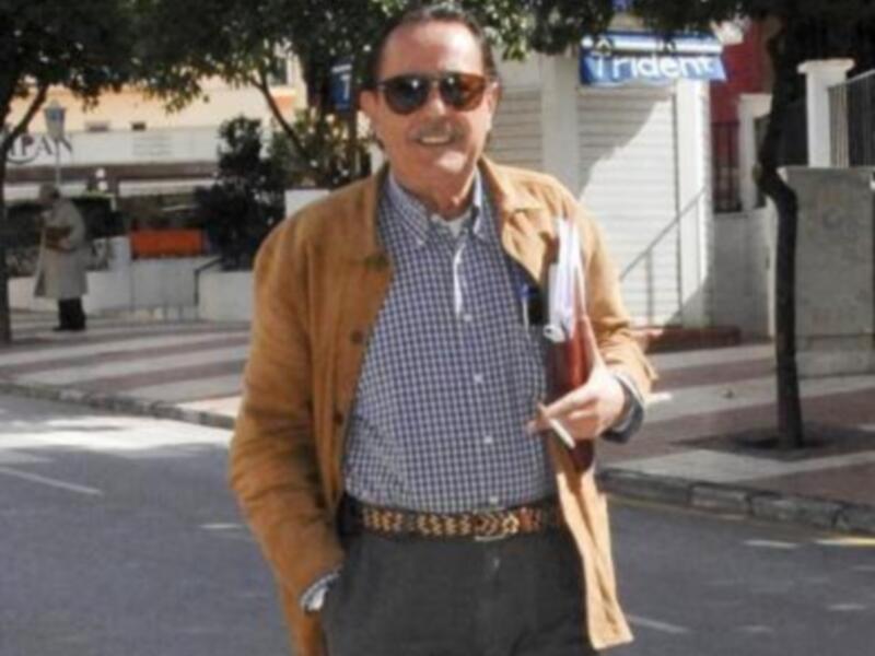 Un hombre con una chaqueta amarilla, camisa a cuadros y pantalones negros camina por una calle con árboles y edificios en el fondo.