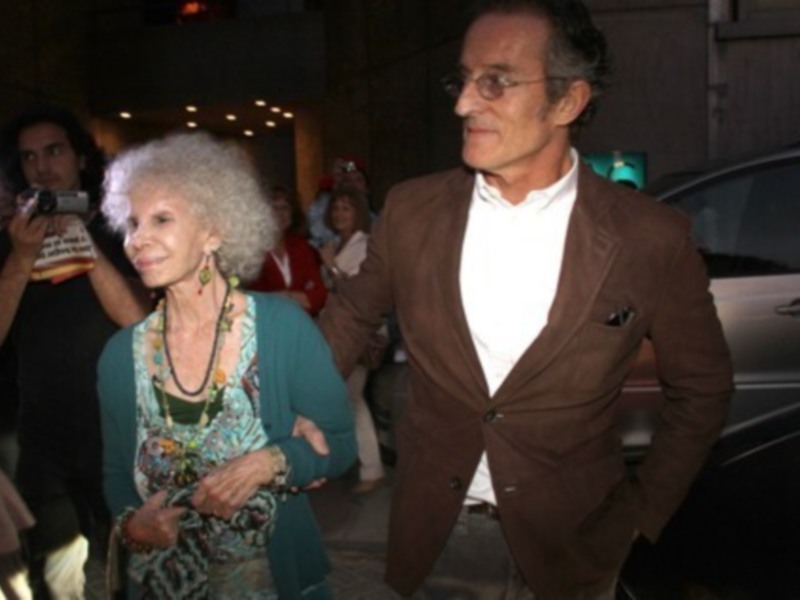 Una anciana con cabello blanco y una mujer joven en el fondo, ambos sonrientes. La anciana lleva un vestido verde con joyería y un collar, mientras que la joven tiene una camisa blanca. En el fondo, hay personas tomando fotos y un coche negro.