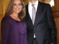 Una pareja elegante posa juntos en un ambiente formal, con una mujer en un vestido morado y un hombre en un traje oscuro.
