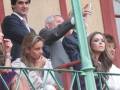 Un grupo de personas en un evento, algunas sonríen y otras observan. La imagen muestra una combinación de personas sentadas en un área de espectadores, con una estructura metálica verde y fondo rojo.