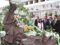 Una estatua de bronce de una mujer con un velo y flores en las manos, rodeada por flores frescas y una multitud de personas en un entierro.