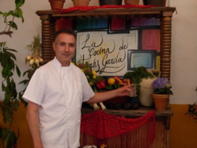 Un hombre en uniforme de chef frente a un mostrador con verduras y flores, detrás hay una pizarra con la escritura "La Cocina de María García".