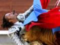 Un torero en traje tradicional se enfrenta a un toro durante una corrida de toros. El torero tiene el rostro cubierto por un tapabocas y lleva una capa azul con detalles dorados. El toro, de color marrón, está en el fondo y parece estar en movimiento.