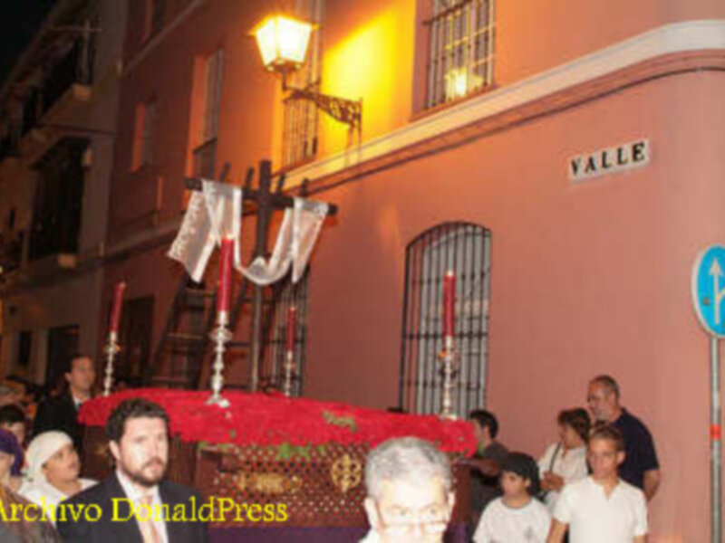 Fotografía nocturna de una procesión religiosa en un barrio con edificios antiguos y farolas. La imagen muestra a personas participando en la procesión, con un paraguas rojo y una farola iluminada. La calle lleva el nombre "Valle".