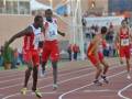Un atleta cubano con camiseta roja y blanco, corriendo en una pista de atletismo. Detrás de él, otros atletas con camisetas rojas y amarillas. En el fondo, una tribuna con espectadores y un cartel que dice "San Fernando".