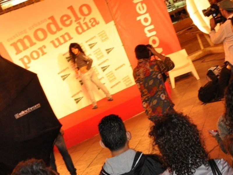 Una modelo posa en un escenario con una pancarta que dice "Modelo por un día". La imagen captura a varias personas observando la modelo, incluyendo a una cámara que filma el evento.