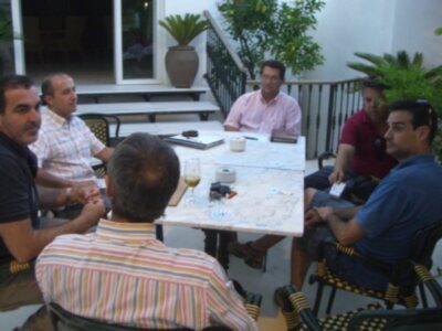 Un grupo de personas se sienta en una mesa al aire libre, disfrutando de un café y charlando. La escena está ambientada en una terraza con plantas verdes y un balcón.