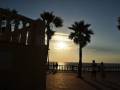 Puesta de sol en un paseo marítimo con palmeras y siluetas de personas caminando.