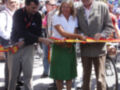 Una mujer corta una cinta roja junto a dos hombres, seguramente en un evento de inauguración o celebración. A su izquierda, hay personas con bicicletas y a la derecha, otros participantes.
