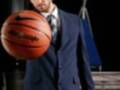 Un hombre con cabello largo y barba, vestido con un traje azul oscuro y corbata, sostiene una pelota de baloncesto Nike. El fondo muestra un aro de baloncesto y una portería, con un logo visible en la parte superior.