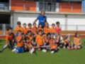 Equipo de fútbol infantil con entrenador en un campo de fútbol artificial bajo el sol.