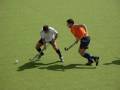 Dos jugadores de hockey en acción sobre un campo verde. Un jugador con camiseta blanca y negro está a punto de golpear una pelota, mientras que el otro jugador con camiseta naranja y azul está corriendo para interceptarla.