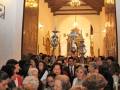 Interior de una iglesia con personas reunidas en un acto religioso. Se destacan dos cruces y una candelabro central. La atmósfera es reverente, con luces doradas y una decoración tradicional.