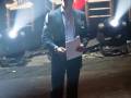 Un hombre en traje negro sostiene un papel mientras se encuentra en una escena de teatro con luces de estudio.