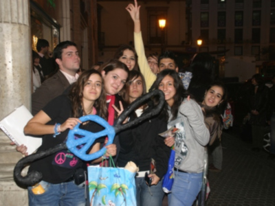 Grupo de jóvenes celebrando con pancarta de paz y bolsas de compras en una calle nocturna.