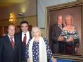 Dos personas posan junto a una pintura de un matrimonio enmarcado. La escena parece ser en un evento formal, posiblemente una recepción o conferencia.