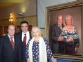 Una familia posa frente a un retrato de una pareja. La imagen muestra a tres personas: dos adultos y una anciana, posando junto al retrato. La escena parece ser en un ambiente formal o ceremonial.