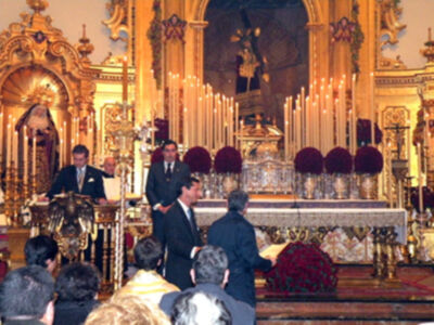 Imagen de una ceremonia religiosa en un templo con personas vestidas formalmente. En el centro, hay una mesa alta decorada con flores y velas encendidas. La arquitectura del templo es elaborada con detalles dorados y oro, creando un ambiente de solemnidad.