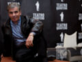 Un hombre sentado en una silla blanca frente a un fondo con el logo del Teatro Quintero, posando con una sonrisa.