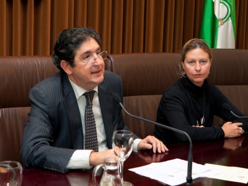 Dos personas sentadas en una mesa de conferencia, uno con traje y corbata, la otra con camiseta negra. En el fondo, una bandera verde y blanca.