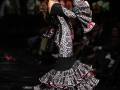 Una modelo desfila con un vestido de flamenca negro y plata, con detalles rojos, mientras sostiene su sombrero. La escena está iluminada y enfocada en la modelo, con un público asistente en el fondo.
