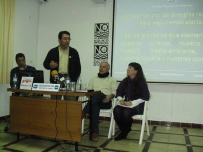 Presentación de un partido político en un auditorio, con tres personas en el escenario y un proyector en el fondo mostrando un mensaje informativo sobre energías renovables y turismo sostenible.