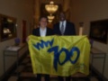 Dos hombres sostienen una bandera amarilla con el texto "WW 700" en letras azules. La imagen se encuentra en un ambiente elegante con una lámpara de techo y una mesa con flores.