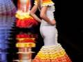 Una modelo presenta un vestido de flamenca con una falda de colores naranja, amarillo y rojo que se extiende hasta el suelo. La falda tiene volantes en la parte inferior, y el vestido es blanco con detalles en rojo y amarillo. La modelo lleva un tocado floral en la cabeza, con flores blancas y hojas verdes. La imagen se enfoca en el detalle del vestido y la expresión de la modelo, que parece estar en movimiento.