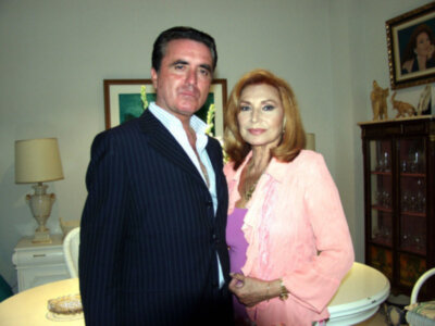A man and a woman standing together in a room with a lamp, a painting, and a cabinet in the background.