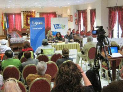 Una sala de conferencias con una mesa central donde se realizan discusiones. En la pared hay un cartel de Cajasol y otro de Cádiz. Hay personas sentadas en sillas rojas, algunos con auriculares y otros con teléfonos. También hay una cámara en un trípode, un portátil y varias banderas colgadas.
