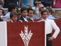 Un grupo de personas detrás de una cerca roja con un símbolo blanco en forma de trigo. Algunos están mirando hacia adelante, mientras otros tienen la vista dirigida hacia el lado. La cerca es de madera y parece estar en un lugar público, posiblemente durante una fiesta o evento.
