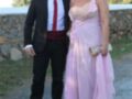Un hombre vestido con traje negro y corbata roja, posando junto a una mujer en un vestido rosa con detalles de encaje y una falda larga. Ambos están en un entorno natural con vegetación y un muro de piedra al fondo.