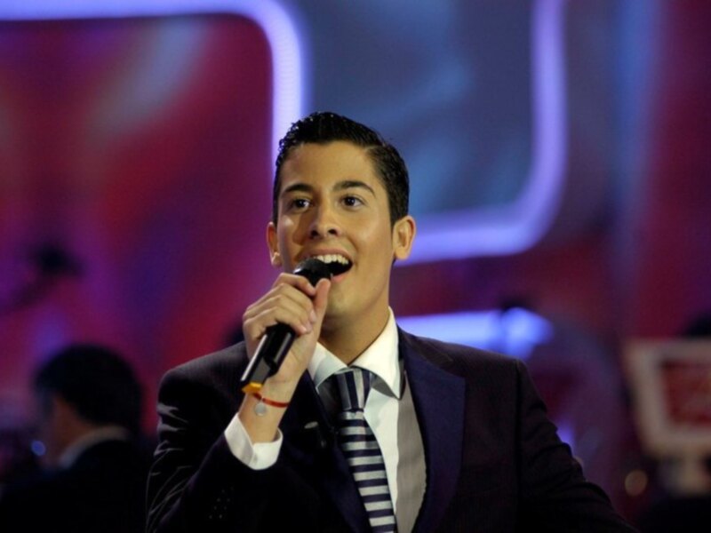 Un hombre en traje y corbata, sosteniendo un micrófono y sonriendo, posiblemente en un evento formal o televisivo.