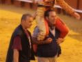 Un torero sonríe mientras es llevado en hombros por un ayudante durante una corrida de toros. El fondo muestra la arena de la plaza de toros con un muro rojo y asientos vacíos.